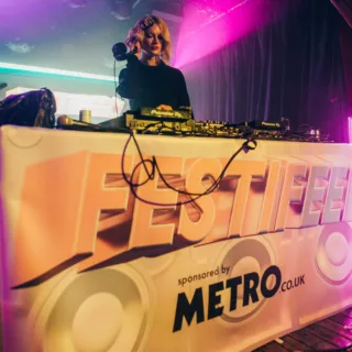 A female DJ performing at a CoppaFeel! Festifeel festival. The DJ decks have Festival branding covering the front and a sponsored by metro.co.uk logo too | CoppaFeel!