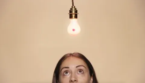 A woman is looking up in thought to a lightbulb that is lite and shaped like a boob