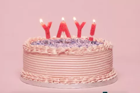 A pink birthday cake decorated in pink piping and purple sprinkles. The cake is topped with pink candles that are lit and read ' yay!'