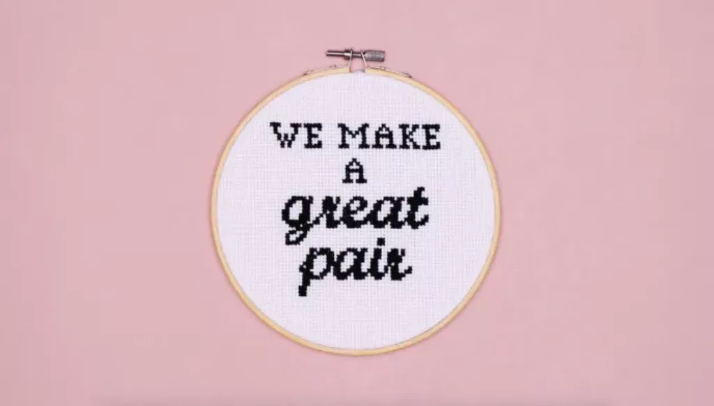 A cross stitch hoop with the wording 'We make a great pair' stitched onto white fabric on a pink background | CoppaFeel!