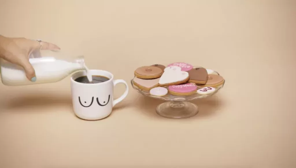 A cup of tea in a white mug with boob illustrations on the front, next to a plate of biscuits decorated with CoppaFeel! branding. A hand is also pouring milk into the cup of tea | CoppaFeel!